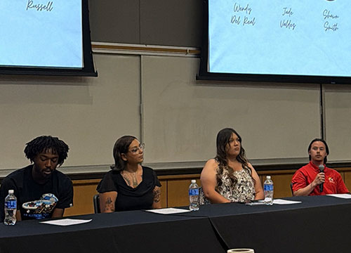 Four student panel members sit at a long table during a presentation in front of projection screens.