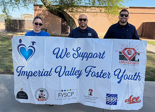 Three campus staff hold a banner reading "We Support Imperial Valley Foster Youth" at an outdoor event.